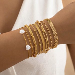 May include: A collection of gold bracelets in various styles adorns a wrist. The bracelets include delicate chains, rope designs, and a mesh band. Two bracelets feature white pearl accents, adding a touch of elegance to the ensemble. The bracelets are displayed against a white background.