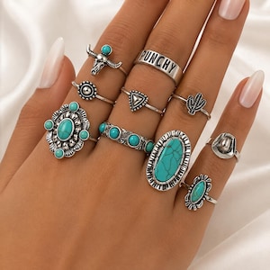 May include: A set of nine silver-toned rings featuring various designs including a bull skull, a band with the text PUNCHY, a cactus, a cowboy hat, and several rings inlaid with oval and round turquoise-colored stones in ornate metal settings.