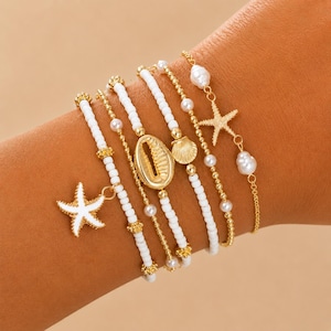 May include: A collection of six bracelets featuring a beach theme. The bracelets include white beaded strands, gold chains, and charms such as starfish, seashells, and pearls. The bracelets are displayed on a light brown surface.