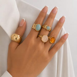 Op de afbeelding: Een hand toont vijf goudkleurige ringen met verschillende edelsteeninstellingen. De ringen hebben een gehamerde textuur, ovale en ronde edelstenen in blauwe, roze en oranje tinten. De achtergrond is wit.