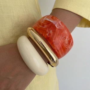 Puede incluir: Tres pulseras apiladas: una pulsera ancha, veteada en naranja y blanco, un tubo rectangular dorado y una pulsera ancha, redondeada y blanquecina. Las pulseras se llevan en una muñeca, sobre un fondo amarillo.