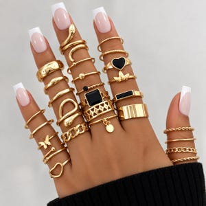 May include: A collection of gold-toned rings worn on fingers, featuring various designs including a snake, a black heart, a black square stone, a butterfly, a chain link, a dangling disc, and smooth bands, all with a polished metallic finish.