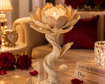 May include: A cream-coloured, flower-shaped candle holder with a lit candle inside. The holder has a twisted stem and sits on a decorative base. Gold accents highlight the edges. Red rose petals and a "For You" card are scattered around.