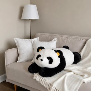 May include: A large, plush panda bear stuffed animal with black and white fur, lying on a beige sofa. The panda has black eyes and ears with yellow accents. Two white pillows and a white blanket with fringe are also on the sofa.