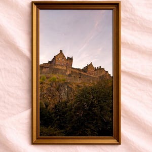 May include: A framed photograph of a castle on a hill, set against a cloudy sky. The castle is made of stone and has multiple towers and walls. The frame is gold-colored, and the photograph is labeled "Heather & Horizon PHOTOGRAPHY".