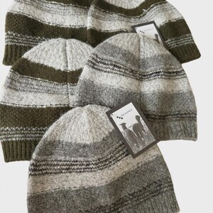 May include: A collection of five knit beanies featuring horizontal stripes in shades of olive green, heather grey, and cream. Each beanie has a ribbed cuff and a small rectangular tag attached, with one tag clearly displaying the text INCALPACA.