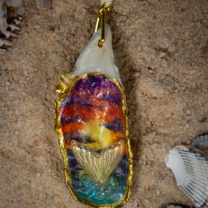 May include: A handcrafted pendant necklace featuring a painted oyster shell with a gold-colored mermaid tail charm. The shell is painted with sunset colors and trimmed with gold. The necklace hangs on a cream-colored cord.