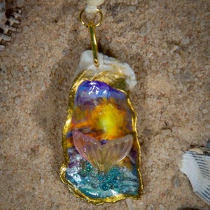 May include: A handcrafted pendant necklace featuring a painted oyster shell with a gold-colored trim. The shell is painted with vibrant colors and a clear, pink mermaid tail charm. The pendant hangs from a cream-colored cord.