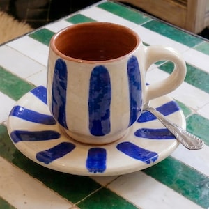 May include: A ceramic cup and saucer set featuring a cream-colored base with hand-painted vertical blue stripes. The cup has a terracotta-colored interior and a sturdy handle. A small silver-toned metal spoon rests on the saucer beside the cup.