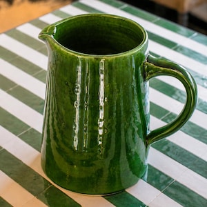 May include: A deep forest green ceramic pitcher with a glossy, reflective glaze. The vessel features a wide, open top with a pronounced pouring spout and a sturdy, curved handle on the side. The surface has a subtle, vertical ribbed texture.