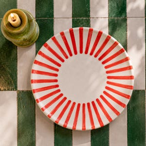 May include: A round ceramic plate featuring a white center and a border of radiating red stripes. Beside the plate sits a small, cylindrical green ceramic candle holder containing a lit cream-colored candle with a visible flame.