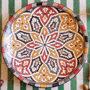 May include: A large circular ceramic platter featuring a complex, symmetrical geometric pattern in vibrant shades of red, yellow, black, and white. The design consists of interlocking star-shaped motifs and intricate floral-inspired shapes.