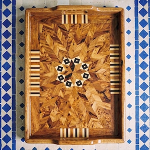 May include: A rectangular wooden serving tray featuring an intricate marquetry design. The surface displays a star-shaped pattern made of light and dark wood veneers, accented by small black square inlays and striped border details on all sides.