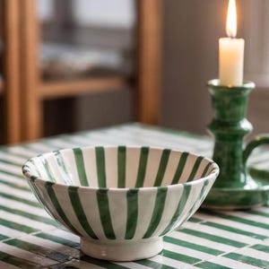 May include: A ceramic bowl featuring a white base with vertical dark green stripes. The bowl has a rounded shape with a raised pedestal base. Beside the bowl stands a green ceramic candle holder containing a lit white taper candle.