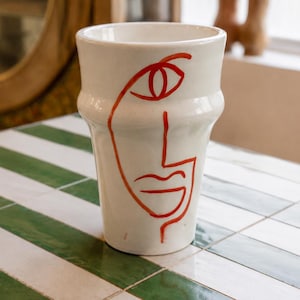 May include: A white ceramic vase with a flared rim and a tapered base, featuring a single continuous line drawing of a human face in bright orange paint. The design includes a stylized eye, a straight nose line, and a curved mouth shape.