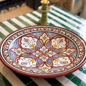 May include: A large, round ceramic platter featuring a hand-painted, intricate geometric and floral pattern in shades of deep red, cobalt blue, yellow, and green on a white background, with a wide, decorative border and a smooth, glazed finish.