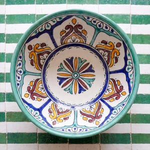 May include: A round ceramic bowl featuring a hand-painted interior with a central floral medallion surrounded by repeating symmetrical patterns in shades of teal, mustard yellow, deep blue, and dark brown on a white background with a teal rim.