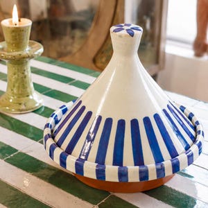May include: A ceramic tagine with a conical lid featuring a white base and vertical blue stripes. The lid is topped with a small knob decorated with a blue floral pattern. The base of the tagine is a solid terracotta color, visible beneath the lid.