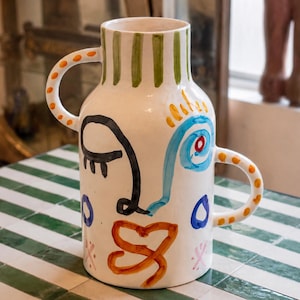 May include: A white ceramic vase with two handles, featuring hand-painted abstract face motifs in black, blue, orange, and green. The neck has vertical green stripes, while the body displays a stylized eye, a swirl, and a knotted orange mouth.