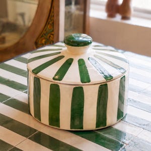 May include: A round ceramic lidded container featuring a cream-colored base with hand-painted dark green vertical stripes. The lid has a matching green knob handle at the center, and the overall design displays a glossy finish on the exterior surface.