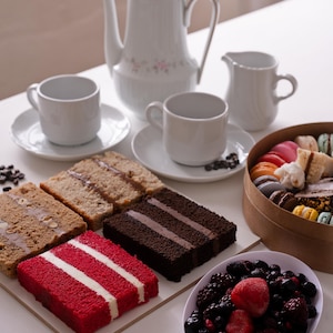 Puede incluir: Un juego de té de porcelana blanca con tetera, crema y dos tazas y platillos. Rebanadas de pastel, incluyendo terciopelo rojo y chocolate, se disponen con un cuenco de bayas y una caja de macarons. Granos de café esparcidos.