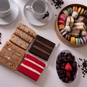 May include: An overhead view of a dessert arrangement with cake slices, a macaron box, and a berry bowl. Two white coffee cups and saucers are at the top, with scattered coffee beans.