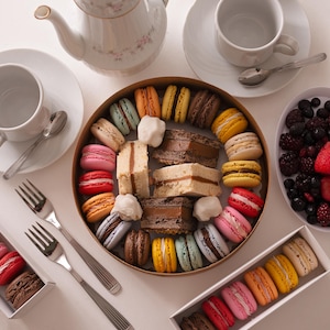 Peut inclure: Une composition de tea party avec une boîte ronde de macarons colorés, des tasses, des soucoupes, des fourchettes, une théière et un bol de baies. Les macarons sont roses, jaunes et bruns.