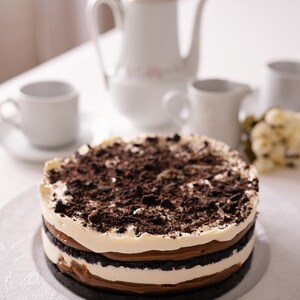 May include: A layered dessert cake with a dark chocolate biscuit base, a creamy white layer, and a chocolate layer, topped with biscuit crumbs. The cake is on a white plate, with a tea set in the background.