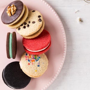 Puede incluir: Un plato rosa contiene una variedad de galletas sándwich coloridas. Las galletas incluyen sabores de chocolate, rojo y vainilla, algunas con chispas y chispas de chocolate. Una nuez cubre una galleta. Las galletas están apiladas.