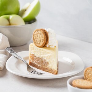 May include: A slice of cheesecake with a layer of apples and a cookie on top, served on a white plate with a fork. Whipped cream is on the side. Green apples are in a bowl in the background.