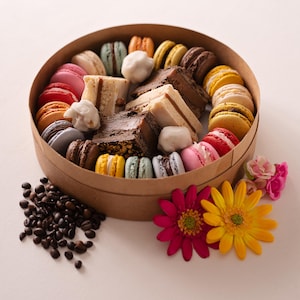 Peut inclure: Une boîte ronde en carton remplie de macarons colorés et de tranches de gâteau. Les macarons sont de différentes couleurs, notamment rose, jaune et bleu. Des grains de café et des fleurs sont disposés autour de la boîte.