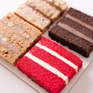 Puede incluir: Una variedad de rebanadas de pastel, incluyendo terciopelo rojo con rayas de glaseado blanco, chocolate con crema de chocolate y otros sabores. Los pasteles están dispuestos sobre una superficie blanca, mostrando sus texturas y capas.
