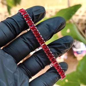 Puede incluir: Una pulsera plateada con gemas rojas rectangulares y vibrantes. Las gemas están muy juntas, creando una línea continua de color. La pulsera es sostenida por una mano enguantada, con un fondo verde borroso.