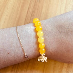 May include: A yellow beaded bracelet with a gold elephant charm. The bracelet features large, round, yellow beads. A delicate gold chain bracelet is also visible.
