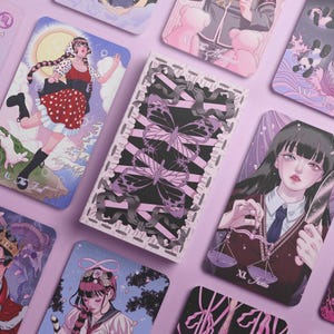May include: A collection of illustrated tarot cards with a pastel color palette. The cards feature various artistic designs, including a woman in a red dress, butterflies, and other figures. The cards are arranged on a light purple surface.