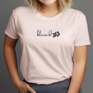 Op de afbeelding: Lichtroze t-shirt met het woord "blessed" in cursief, vergezeld van een zwart bloemenontwerp. Het shirt heeft korte mouwen en is gemaakt van een zacht materiaal.
