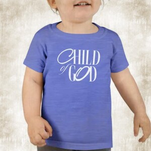 Op de afbeelding: Een blauw T-shirt met de woorden "CHILD of GOD" in witte letters. Het shirt met korte mouwen lijkt van een zachte stof te zijn. De afbeelding toont het shirt op een kind.