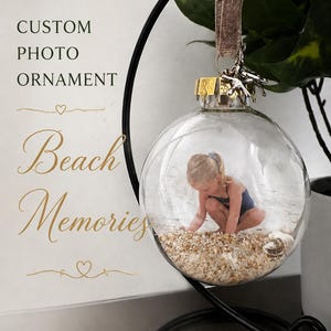 May include: Clear glass ornament with a photo of a child playing on a beach, filled with sand and seashells. The ornament hangs from a black metal stand. Text on the image reads "Custom Photo Ornament" and "Beach Memories".