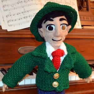 Puede incluir: Una muñeca de crochet que lleva una chaqueta verde, pantalones azules y un sombrero verde con una corbata roja. La muñeca está de pie sobre una superficie de madera con un teclado en el fondo.