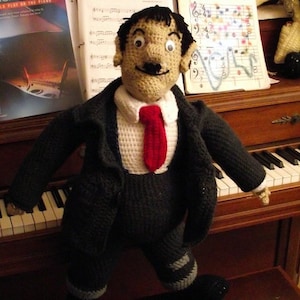 May include: A crocheted doll wearing a black suit and a red tie. The doll is standing on a piano keyboard with a black hat on the floor.