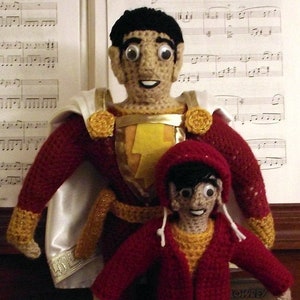 May include: Two crocheted dolls, one dressed as a superhero with a red cape and yellow lightning bolt, the other in a red hooded sweater and blue jeans, standing on a piano keyboard.