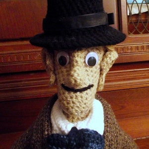 May include: A crocheted doll wearing a black fedora hat, a white collared shirt, a brown sweater, and a blue bow tie. The doll has large, round eyes and a crocheted face.