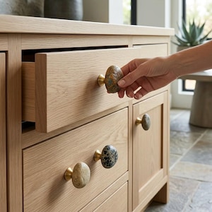 May include: A light oak wood cabinet featuring several drawer and door pulls made from natural, smooth, oval-shaped stones attached to brass-coloured metal bases. The stones vary in colour, including shades of tan, grey, and speckled brown.