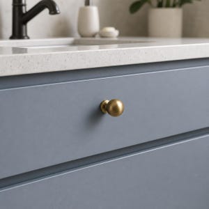 May include: A round, brushed brass cabinet knob with a smooth, spherical shape and a metallic finish, mounted on a flat, matte blue drawer front. The knob features a simple, circular base that connects directly to the drawer surface.