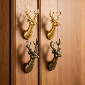 May include: Four metal cabinet knobs shaped like deer heads, arranged in a two-by-two grid. The top two knobs have a polished, bright gold finish, while the bottom two knobs feature a darker, antique brass finish with visible textured detailing.