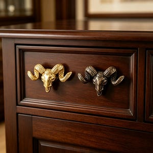 Op de afbeelding: Twee metalen ladeknoppen in de vorm van ramschedels zijn gemonteerd op een donkerhouten lade. De ene knop heeft een gepolijste gouden afwerking, terwijl de andere een donkere, geantiekte bronzen afwerking heeft. Beide tonen gedetailleerde hoorns.