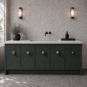 Puede incluir: Un mueble de baño verde oscuro con cinco puertas de armario, cada una con un pomo redondo de latón. El mueble tiene una encimera de piedra gris claro, un grifo negro y una bandeja negra con un dispensador de jabón negro encima.