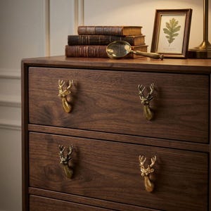 May include: A dark brown wooden dresser featuring four drawers, each equipped with a gold-toned metal deer head handle. The dresser surface holds a stack of three vintage books, a gold-toned magnifying glass, and a framed botanical leaf print.