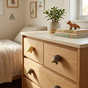 Puede incluir: Una cómoda de madera de roble claro con cuatro cajones y tiradores metálicos en forma de erizo. La superficie superior sostiene una planta verde en una maceta de cerámica y una pila de dos libros coronada por una pequeña figura de zorro.