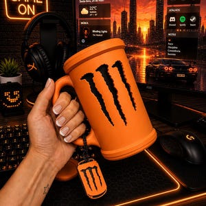 May include: A bright orange mug featuring a cutout of the three-claw Monster Energy logo on its side. The mug includes a matching orange lid and a small, dangling orange dog tag-style charm that also displays the black three-claw logo.